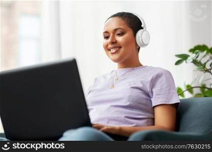 people, technology and leisure concept - happy young african american woman in headphones with laptop computer listening to music at home. woman with laptop listening to music at home