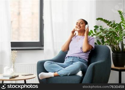 people, technology and leisure concept - happy young african american woman in headphones sitting in chair and listening to music at home. woman in headphones listening to music at home