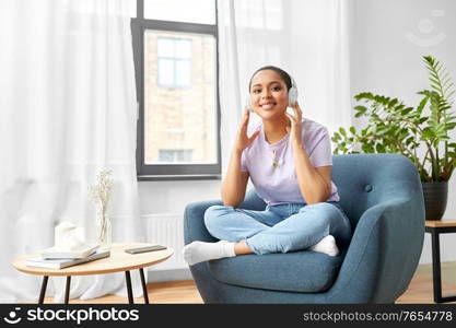 people, technology and leisure concept - happy young african american woman in headphones sitting in chair and listening to music at home. woman in headphones listening to music at home