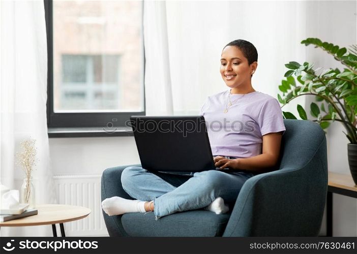 people, technology and leisure concept - happy young african american woman sitting in chair with laptop computer at home. african american woman with laptop at home