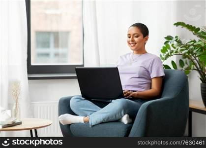 people, technology and leisure concept - happy young african american woman sitting in chair with laptop computer at home. african american woman with laptop at home