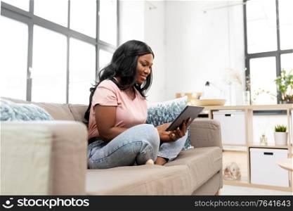 people, technology and leisure concept - happy young african american woman sitting on sofa with tablet pc computer at home. african american woman with tablet pc at home