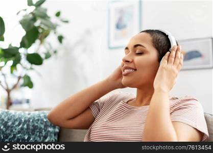 people, technology and leisure concept - happy young african american woman with headphones sitting on sofa and listening to music at home. woman in headphones listening to music at home