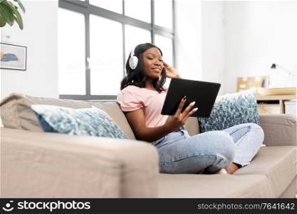 people, technology and leisure concept - happy young african american woman with tablet pc computer and headphones sitting on sofa and listening to music at home. woman with tablet pc listening to music at home