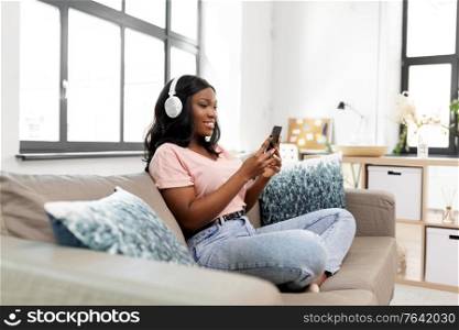 people, technology and leisure concept - happy young african american woman with smartphone and headphones sitting on sofa and listening to music at home. woman with smartphone listening to music at home