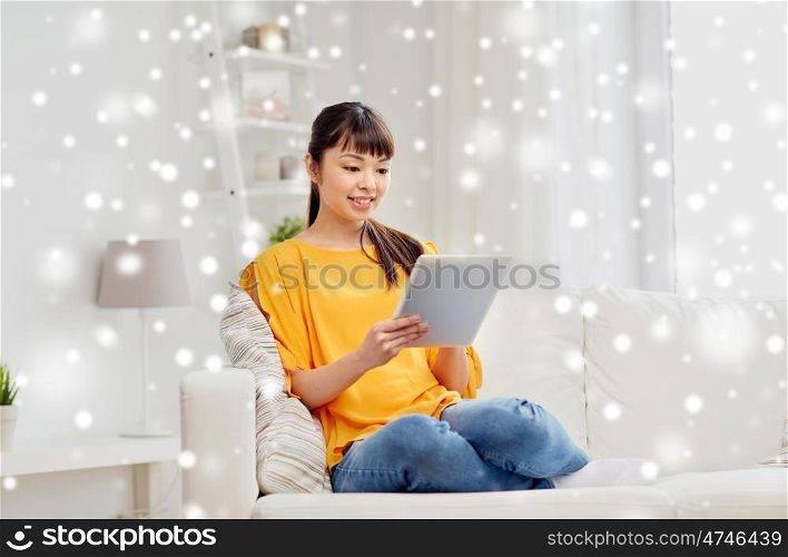 people, technology and leisure concept - happy young asian woman sitting on sofa with tablet pc computer at home over snow