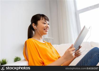 people, technology and leisure concept - happy young asian woman sitting on sofa in earphones with tablet pc computer and listening to music at home. happy asian woman with tablet pc and earphones
