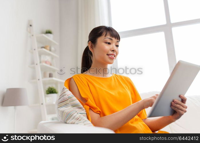 people, technology and leisure concept - happy young asian woman sitting on sofa with tablet pc computer at home. happy young asian woman with tablet pc at home