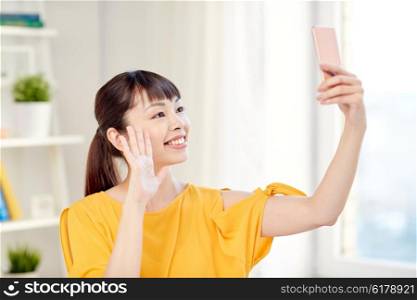 people, technology and leisure concept - happy young asian woman taking selfie on smartphone at home and waving hand