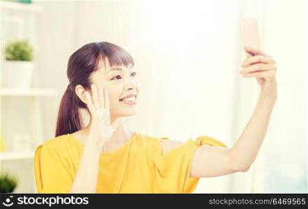 people, technology and leisure concept - happy young asian woman taking selfie on smartphone at home and waving hand. happy asian woman taking selfie with smartphone