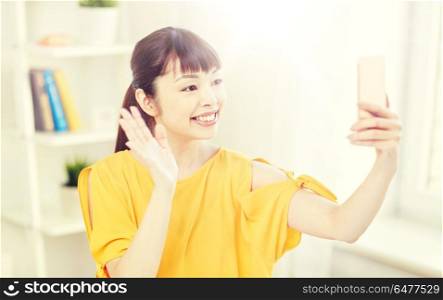 people, technology and leisure concept - happy young asian woman taking selfie on smartphone at home and waving hand. happy asian woman taking selfie with smartphone. happy asian woman taking selfie with smartphone