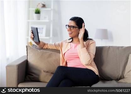 people, technology and leisure concept - happy young indian woman sitting on sofa with tablet pc computer and having video call at home. woman with tablet pc having video call at home
