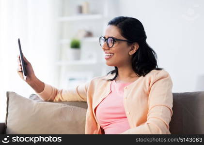 people, technology and leisure concept - happy young indian woman sitting on sofa with tablet pc computer at home. happy indian woman with tablet pc at home