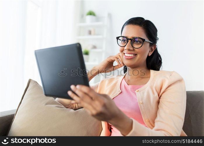 people, technology and leisure concept - happy young indian woman sitting on sofa with tablet pc computer at home. happy indian woman with tablet pc at home