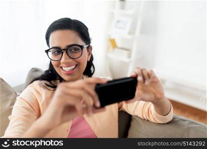 people, technology and leisure concept - happy young indian woman taking selfie on smartphone at home. happy woman taking selfie with smartphone at home