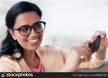 people, technology and leisure concept - happy young woman in glasses taking selfie or photographing on smartphone at home. happy woman taking selfie with smartphone at home