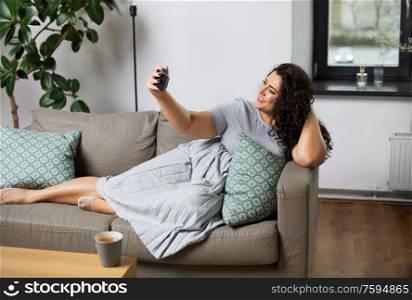 people, technology and leisure concept - happy young woman lying on sofa and taking selfie with smartphone at home. happy woman taking selfie with smartphone at home