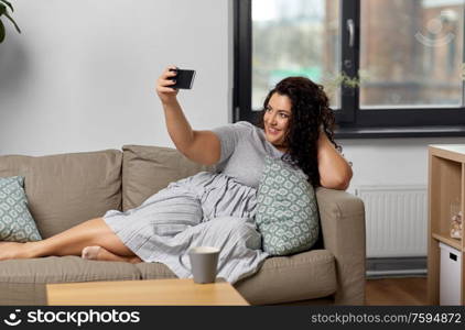 people, technology and leisure concept - happy young woman lying on sofa and taking selfie with smartphone at home. happy woman taking selfie with smartphone at home