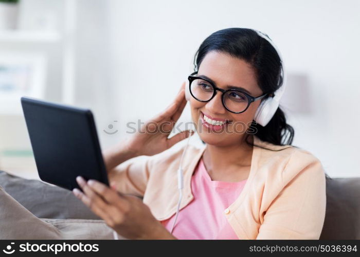 people, technology and leisure concept - happy young woman sitting on sofa with tablet pc computer and headphones listening to music at home. happy woman with tablet pc and headphones at home