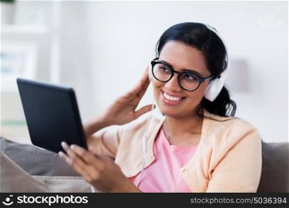 people, technology and leisure concept - happy young woman sitting on sofa with tablet pc computer and headphones listening to music at home. happy woman with tablet pc and headphones at home