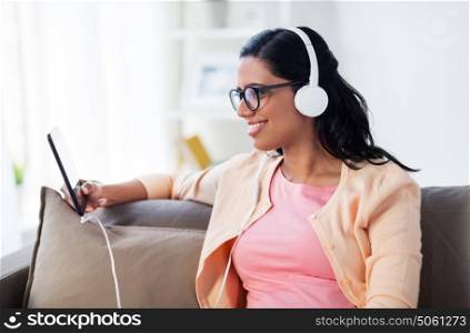 people, technology and leisure concept - happy young woman sitting on sofa with tablet pc computer and headphones listening to music at home. happy woman with tablet pc and headphones at home