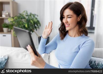 people, technology and leisure concept - happy young woman sitting on sofa with tablet pc computer having video call and waving hand at home. woman having video call on tablet pc at home