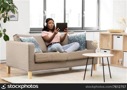 people, technology and leisure concept - surprised young african american woman with tablet pc computer and headphones sitting on sofa and listening to music at home. woman with tablet pc listening to music at home