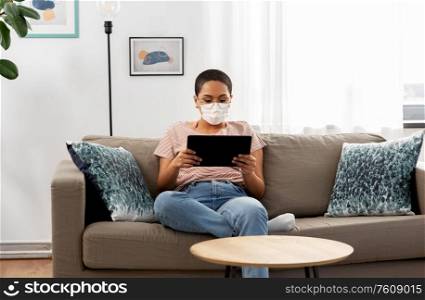 people, technology and leisure concept - young african american woman in glasses and face protective medical mask for protection from virus disease sitting on sofa with tablet pc computer at home. woman in medical mask with tablet pc at home
