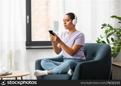 people, technology and leisure concept - young african american woman in glasses with smartphone and headphones sitting in chair and listening to music at home. woman with smartphone listening to music at home