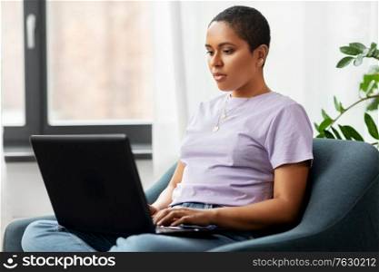 people, technology and leisure concept - young african american woman sitting in chair with laptop computer at home. african american woman with laptop at home