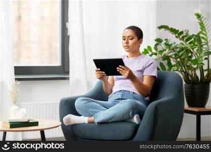 people, technology and leisure concept - young african american woman with tablet pc computer sitting in chair at home. african american woman with tablet pc at home