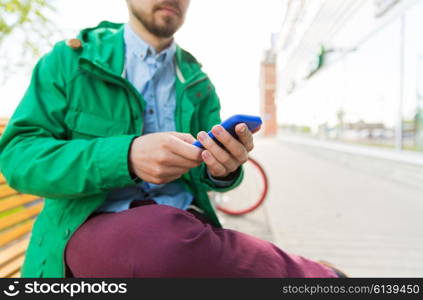 people, technology and lifestyle - close up of young hipster man with smartphone and sitting on street bench in city