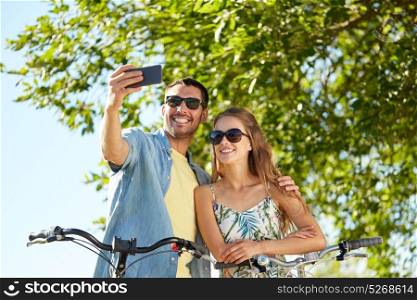 people, technology and lifestyle concept - happy couple with bicycles taking selfie by smartphone at country in summer. couple with bicycles taking selfie by smartphone