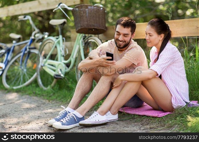 people, technology and lifestyle concept - happy couple with smartphone and bicycles at summer park. couple with smartphone and bicycles at summer park