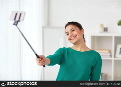 people, technology and lifestyle concept - happy woman taking picture with smartphone selfie stick or monopod at home. woman taking selfie by smartphone monopod at home