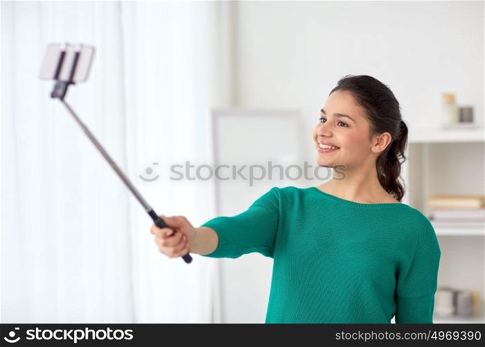 people, technology and lifestyle concept - happy woman taking picture with smartphone selfie stick or monopod at home. woman taking selfie by smartphone monopod at home