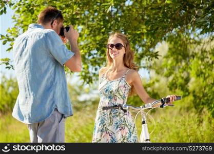 people, technology and lifestyle concept - man photographing happy woman with bicycle by camera at country in summer. man photographing woman with bicycle by camera