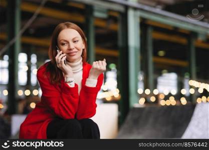 People, technology and lifestyle concept. Positive Caucasian woman has telephone conversation, talks to friends via cellular, wears red coat and black trousers, poses outside with free space