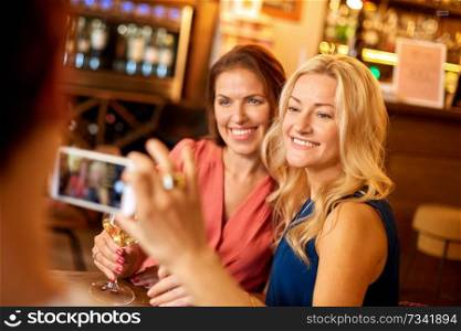 people, technology and lifestyle concept - woman photographing friends by smartphone at wine bar or restaurant. woman picturing friends by smartphone at wine bar