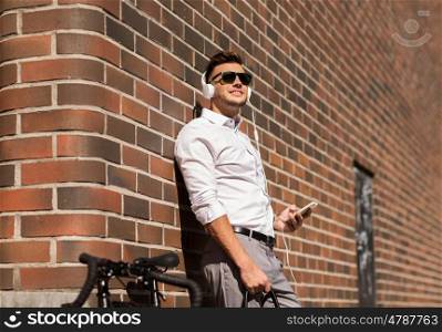 people, technology and lifestyle - happy young man with headphones, smartphone and bicycle listening to music in city