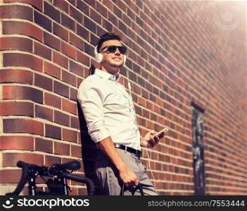 people, technology and lifestyle - happy young man with headphones, smartphone and bicycle listening to music in city. man with headphones, smartphone and bicycle