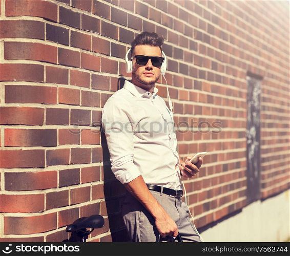 people, technology and lifestyle - young man with headphones, smartphone and bicycle listening to music in city. man with headphones, smartphone and bicycle