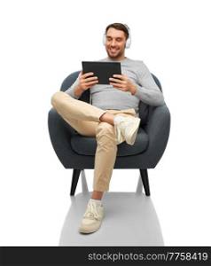 people, technology and music concept - happy smiling man with headphones and tablet pc computer sitting in chair over white background. man with headphones and tablet pc sitting in chair