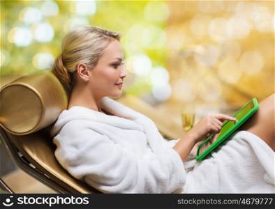 people, technology and relaxation concept - beautiful young woman in white bath robe with tablet pc computer social networking at spa over holidays lights background
