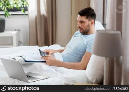 people, technology and remote job concept - man with calculator, papers and laptop computer working in bed at home bedroom. man with calculator, bills and laptop works in bed