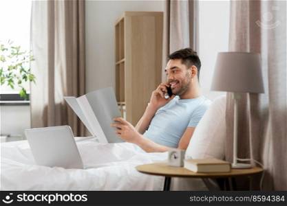 people, technology and remote job concept - man with folder and laptop computer calling on smartphone in bed at home bedroom. man with folder calling on phone in bed at home