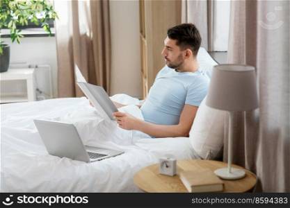 people, technology and remote job concept - man with laptop computer and folder working in bed at home bedroom. man with laptop working in bed at home bedroom