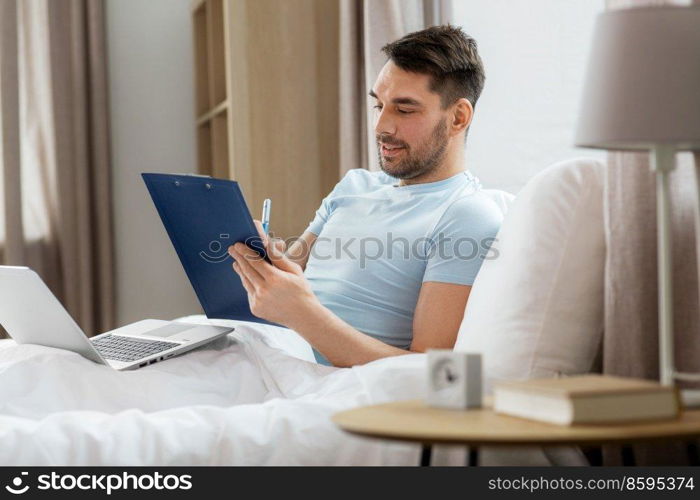 people, technology and remote job concept - man with laptop computer and clipboard working in bed at home bedroom. man with laptop working in bed at home bedroom