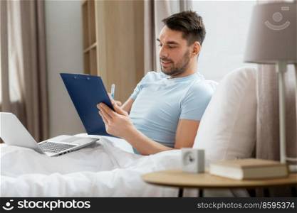 people, technology and remote job concept - man with laptop computer and clipboard working in bed at home bedroom. man with laptop working in bed at home bedroom