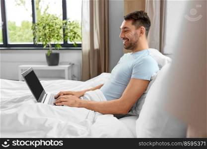 people, technology and remote job concept - man with laptop computer lying in bed at home bedroom. man with laptop in bed at home bedroom